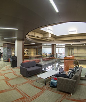 A student sitting on a couch, working on a computer in an indoor space on campus.